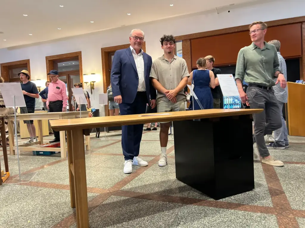 Gabriel Ferreira Marques_Gesellenstueck_Albani_28 Ein Holztisch mit schwarzem Untergestell, präsentiert von zwei Personen in einem Raum mit Publikum.